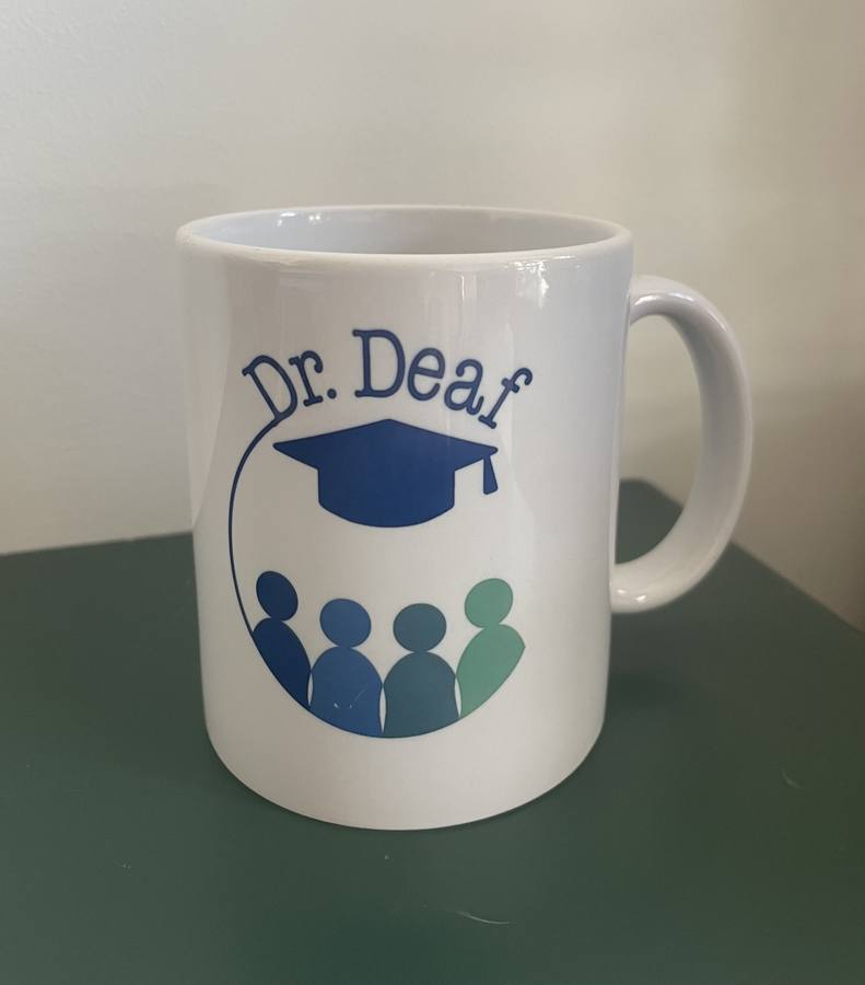 The white Dr. Deaf mug on a dark green surface, showing the full logo